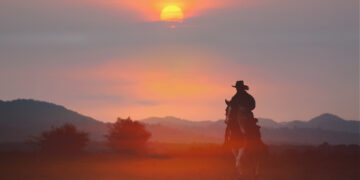 Silhouette of a rider on a horse at sunset, with a warm orange sky and distant hills behind.