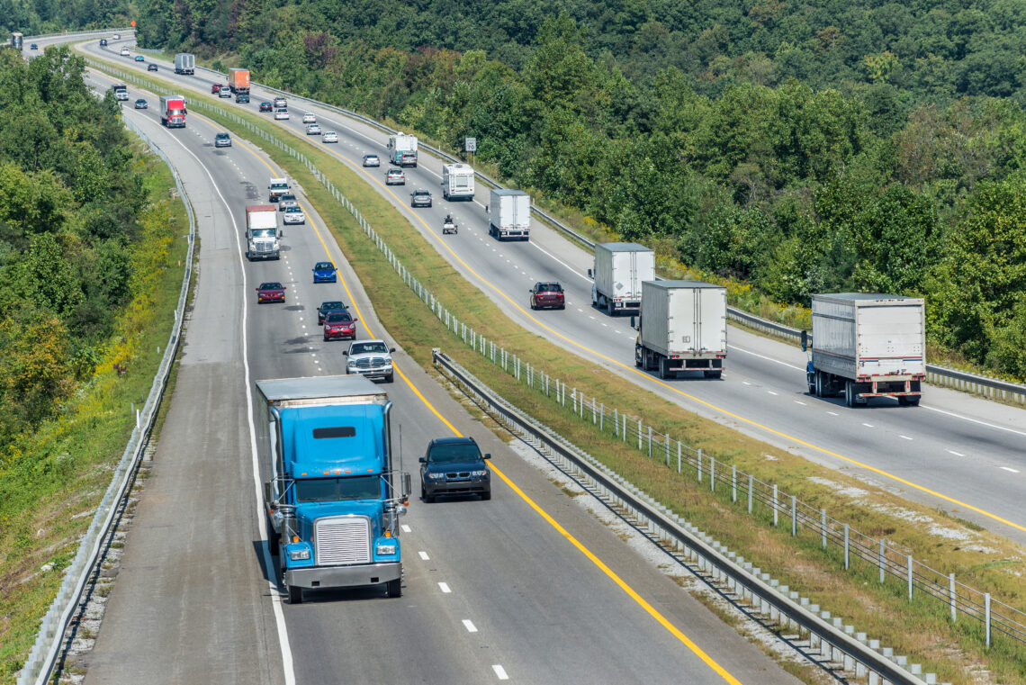 Wide aerial view of a multi-lane highway flanked by dense trees, with several trucks and cars traveling in both directions on a sunny day.