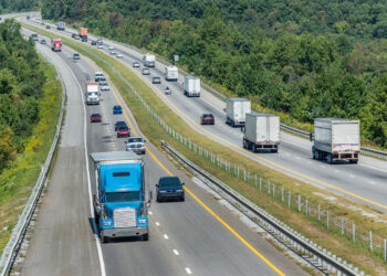 Wide aerial view of a multi-lane highway flanked by dense trees, with several trucks and cars traveling in both directions on a sunny day.