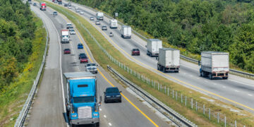 Wide aerial view of a multi-lane highway flanked by dense trees, with several trucks and cars traveling in both directions on a sunny day.