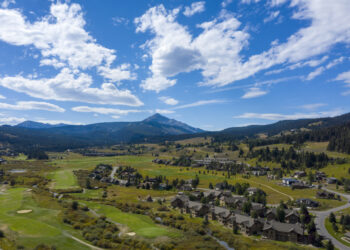 Aerial view of a hillside town with houses, green golf courses, and distant mountains under a blue cloudy sky.