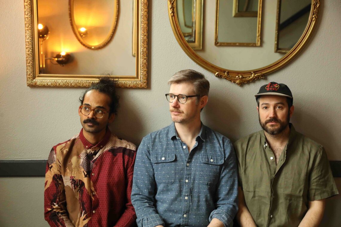 Three men sit against a wall with gold-framed mirrors above; left wears a red patterned shirt, middle wears a blue check shirt, right wears an olive shirt and hat.