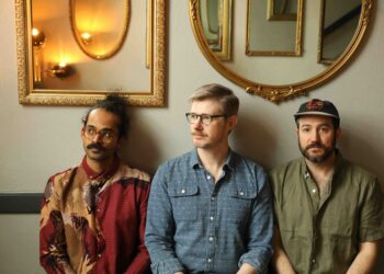 Three men sit against a wall with gold-framed mirrors above; left wears a red patterned shirt, middle wears a blue check shirt, right wears an olive shirt and hat.