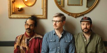 Three men sit against a wall with gold-framed mirrors above; left wears a red patterned shirt, middle wears a blue check shirt, right wears an olive shirt and hat.