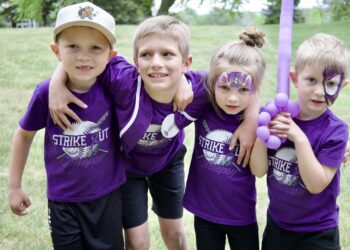 Four children in matching purple shirts stand close together outdoors, some with face paint, smiles for a group photo with a purple balloon nearby.