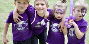 Four children in matching purple shirts stand close together outdoors, some with face paint, smiles for a group photo with a purple balloon nearby.