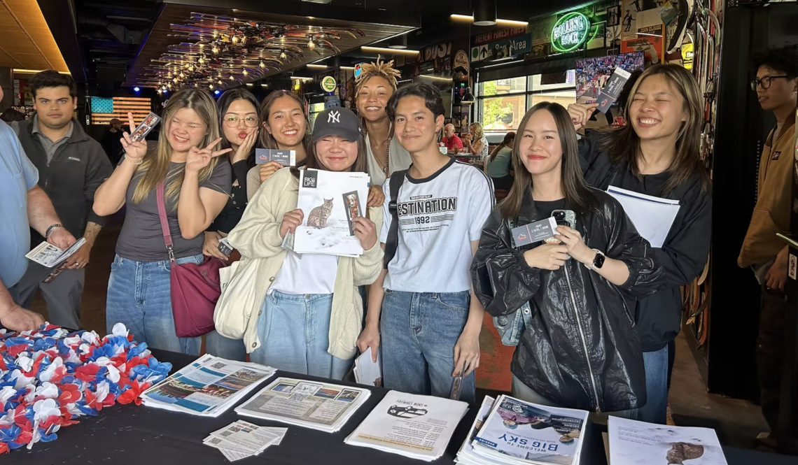 Group of diverse people smiling and posing for a photo at a booth, holding flyers and newsletters inside a busy shop or venue interior.