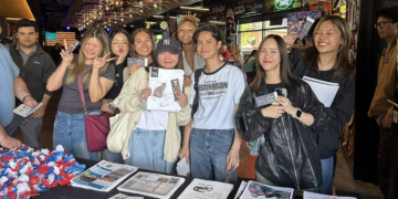 Group of diverse people smiling and posing for a photo at a booth, holding flyers and newsletters inside a busy shop or venue interior.