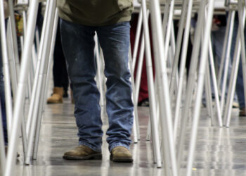 Person in blue jeans and brown boots walking between rows of folding tables with metal legs.