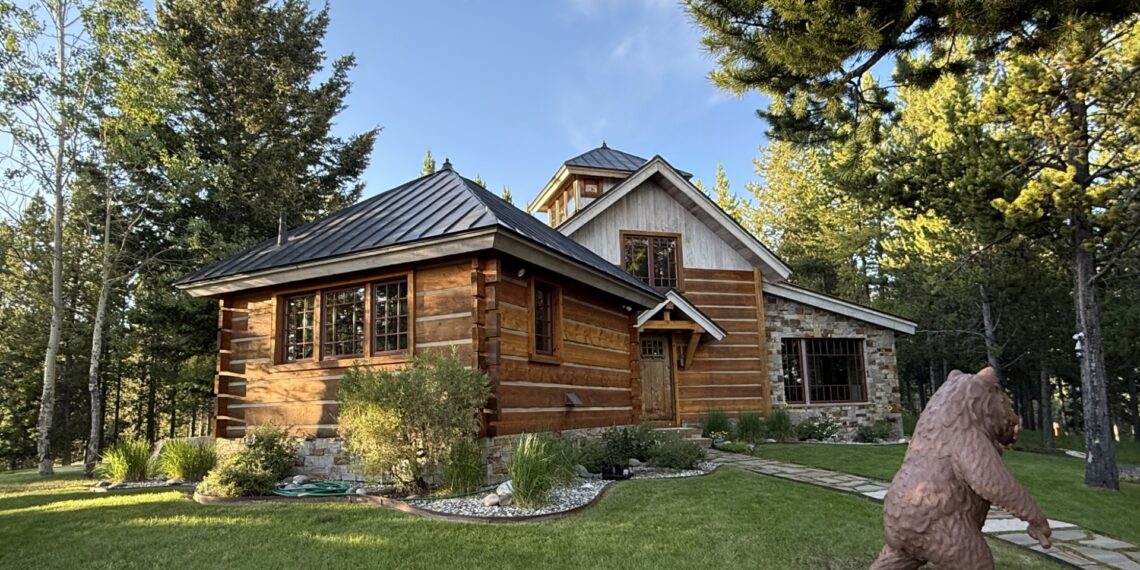 Log cabin with a stone foundation in a wooded yard; a large bear statue is in the foreground on the right side of the yard.