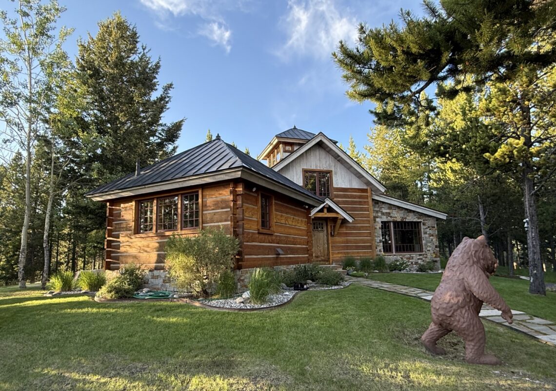 Log cabin with a stone foundation in a wooded yard; a large bear statue is in the foreground on the right side of the yard.