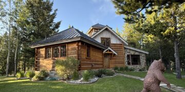 Log cabin with a stone foundation in a wooded yard; a large bear statue is in the foreground on the right side of the yard.