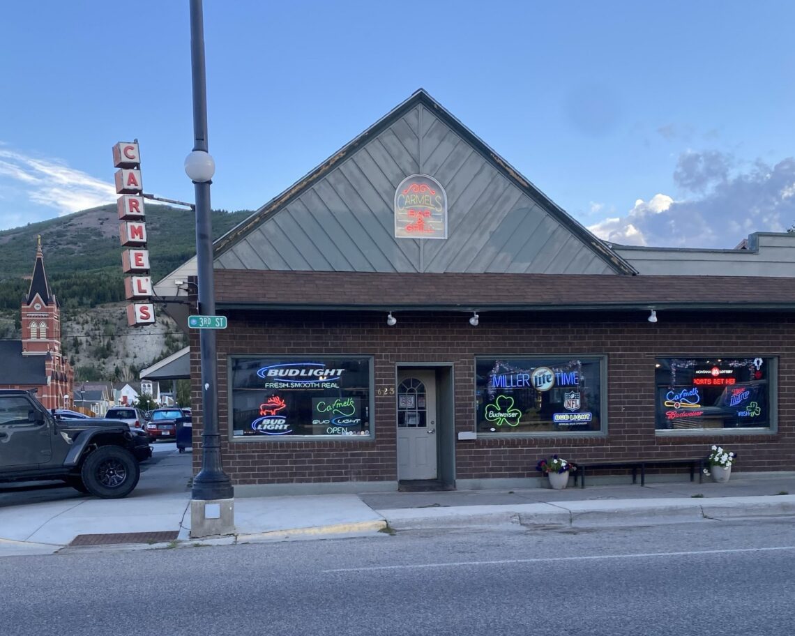 Front view of Carmel's Bar & Grill building with neon signs in the windows and a tall vertical 'CARMEL'S' sign on the left against a blue sky.