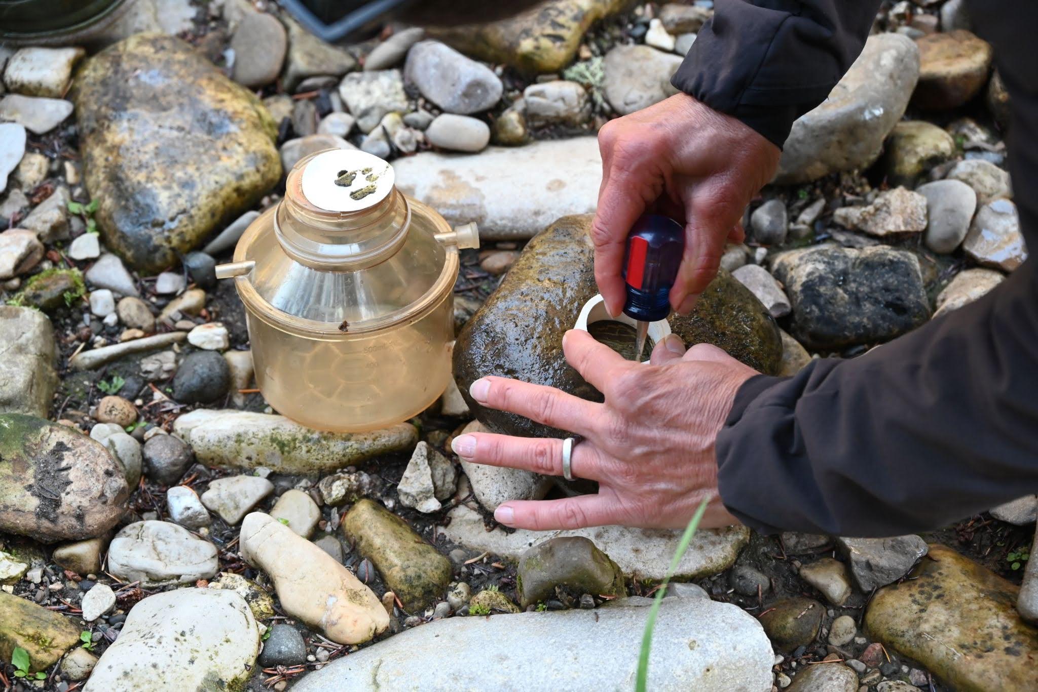 Person outdoors on a rocky shore uses a dropper to add liquid to a large clear jar with measurement marks.