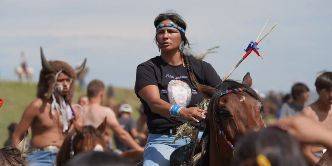 Native American woman rides a horse at an outdoor gathering, wearing a blue patterned headband and bead earrings with a crowd in the background.