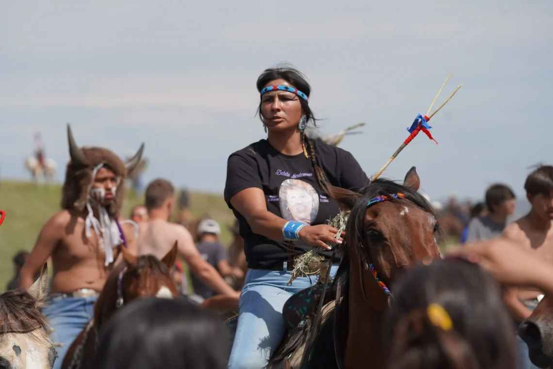 Native American woman rides a horse at an outdoor gathering, wearing a blue patterned headband and bead earrings with a crowd in the background.