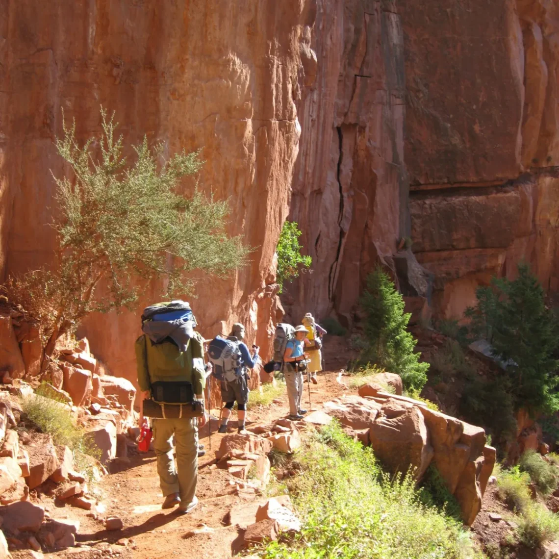Group of hikers with large backpacks walking along a rocky desert trail beside a red cliff wall.