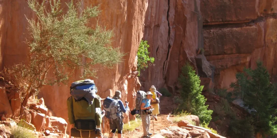Group of hikers with large backpacks walking along a rocky desert trail beside a red cliff wall.