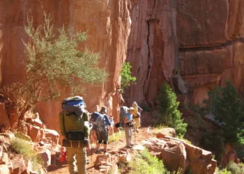 Group of hikers with large backpacks walking along a rocky desert trail beside a red cliff wall.