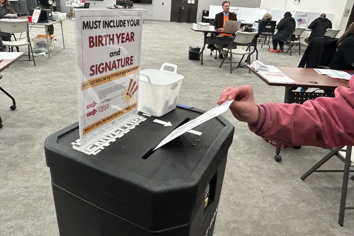 Voter inserts a ballot into a ballot drop box at a polling station.”
