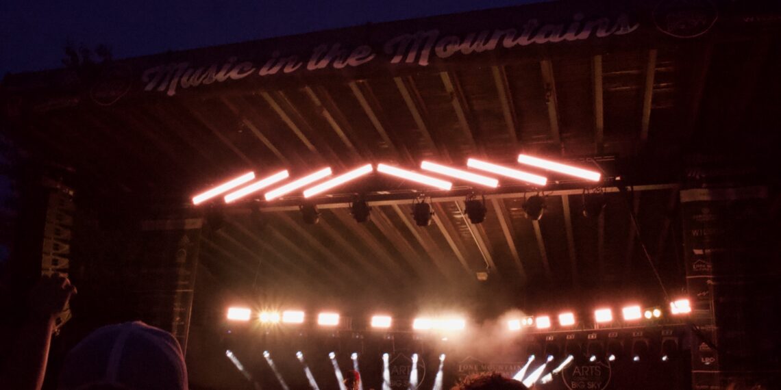 Nighttime outdoor concert with a performer on a lit stage and a silhouetted crowd in the foreground. Neon strip lights run diagonally above the stage.