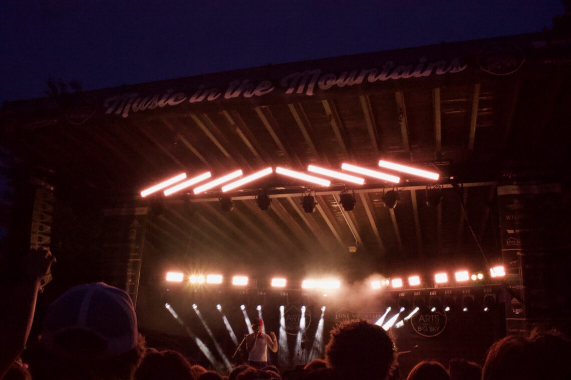 Nighttime outdoor concert with a performer on a lit stage and a silhouetted crowd in the foreground. Neon strip lights run diagonally above the stage.