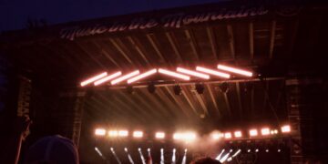 Nighttime outdoor concert with a performer on a lit stage and a silhouetted crowd in the foreground. Neon strip lights run diagonally above the stage.