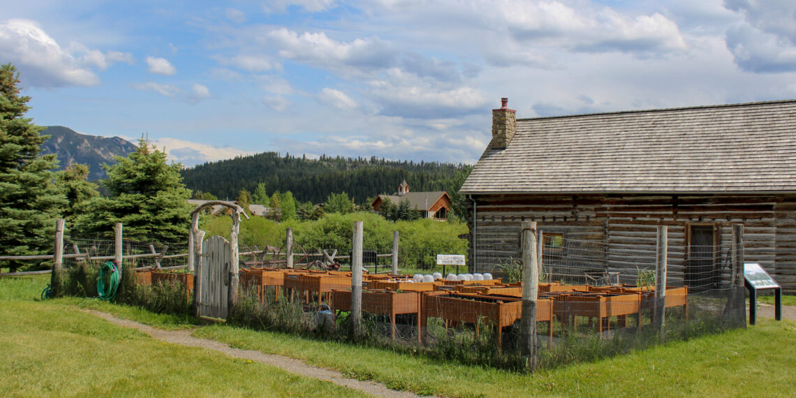 Rural farm scene with a large wooden barn, stacked orange crates, and a grassy yard, mountains in the distance under a blue cloudy sky.