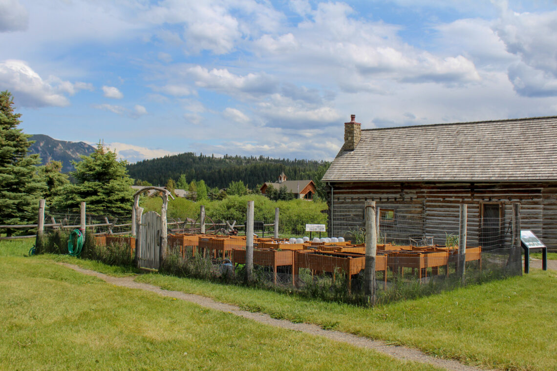 Rural farm scene with a large wooden barn, stacked orange crates, and a grassy yard, mountains in the distance under a blue cloudy sky.