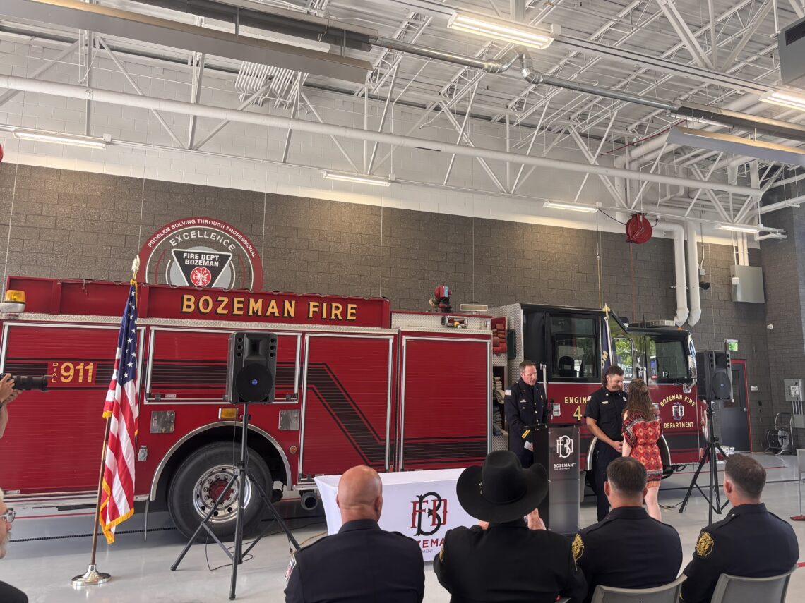 Inside Bozeman Fire Department hall with a red fire engine, flags, and seated audience near a stage ceremony area.