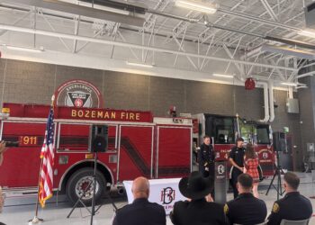 Inside Bozeman Fire Department hall with a red fire engine, flags, and seated audience near a stage ceremony area.
