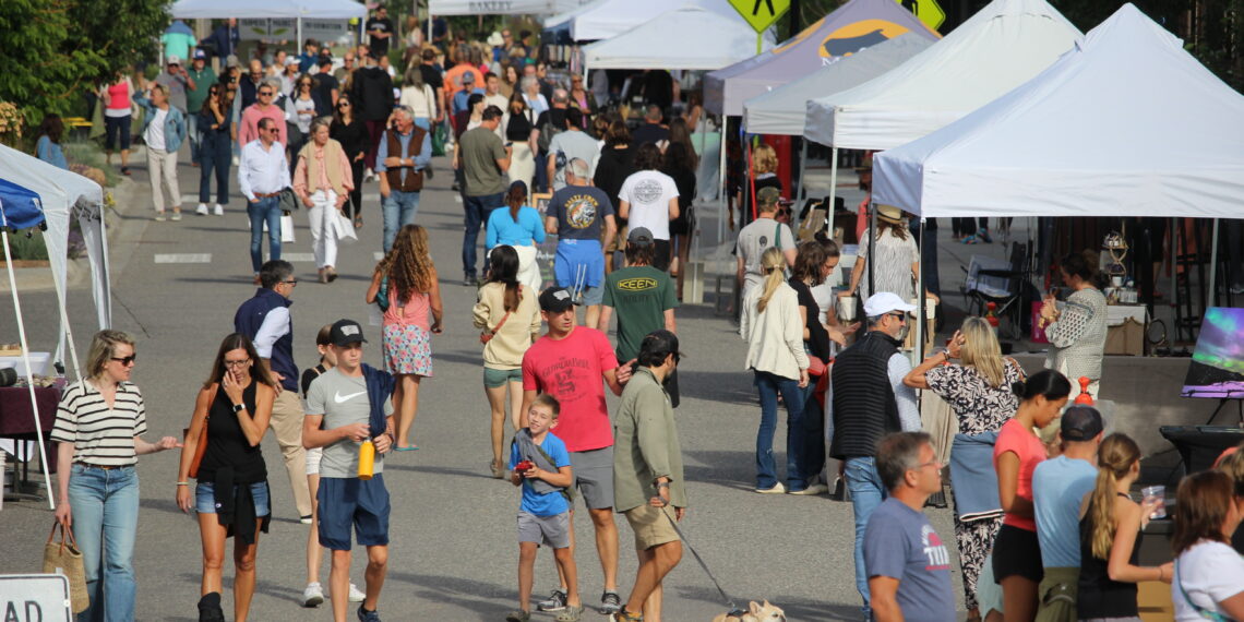 Crowd of people walking between vendor tents at a daylight street fair; a small dog on a leash in the foreground.