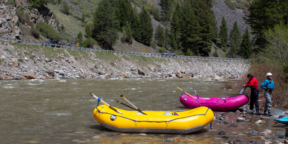 Rafts by a rocky riverbank with people in life jackets, a bright yellow raft in the foreground, and a forested hillside in the background.