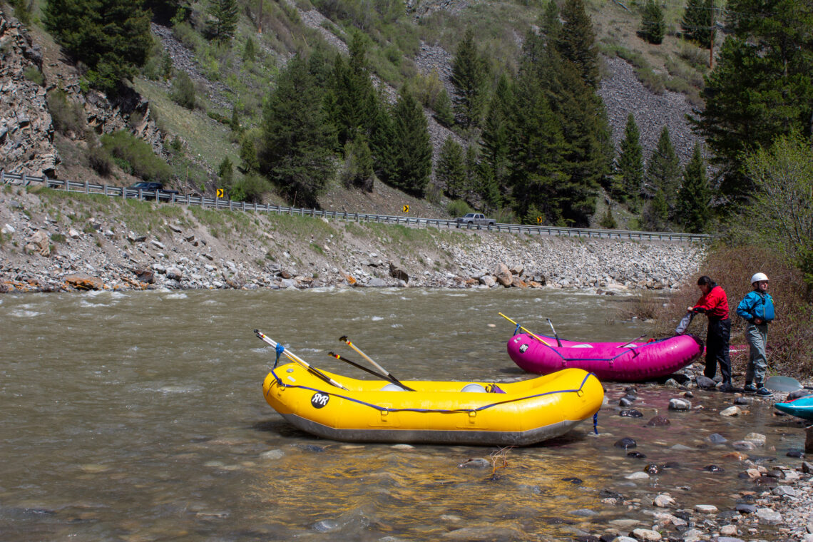 Rafts by a rocky riverbank with people in life jackets, a bright yellow raft in the foreground, and a forested hillside in the background.