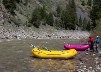 Rafts by a rocky riverbank with people in life jackets, a bright yellow raft in the foreground, and a forested hillside in the background.