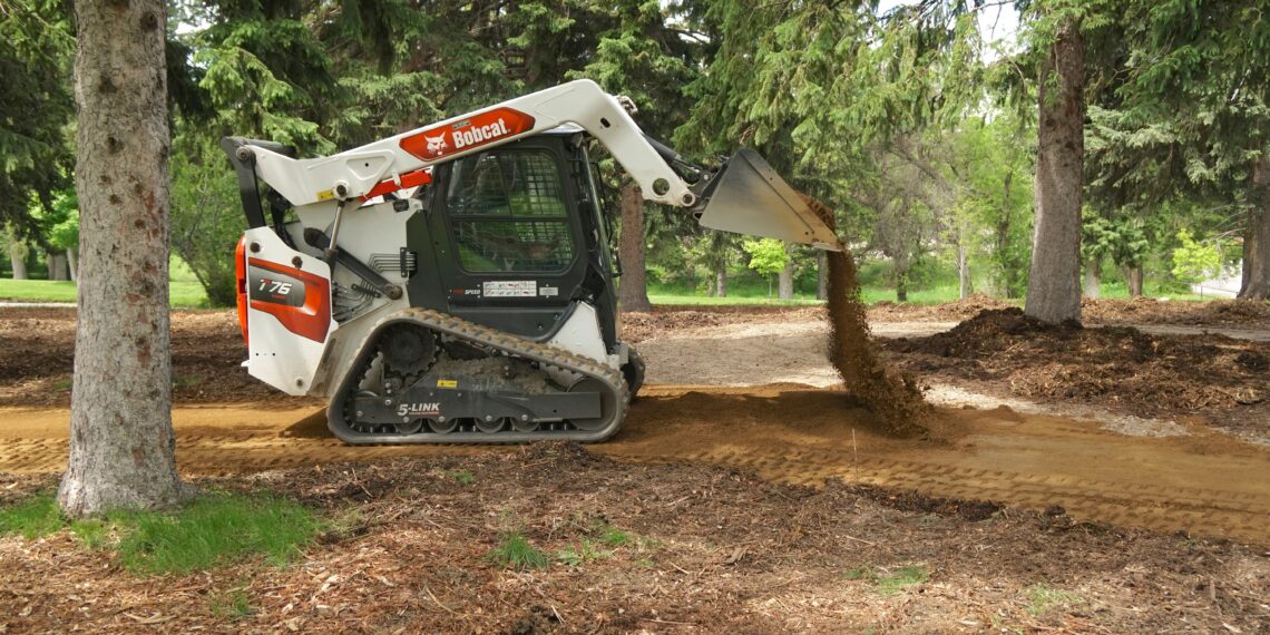 Bobcat skid-steer loader dumping soil in a wooded park, dirt pouring from bucket onto the ground amidst trees and mulch-covered soil.