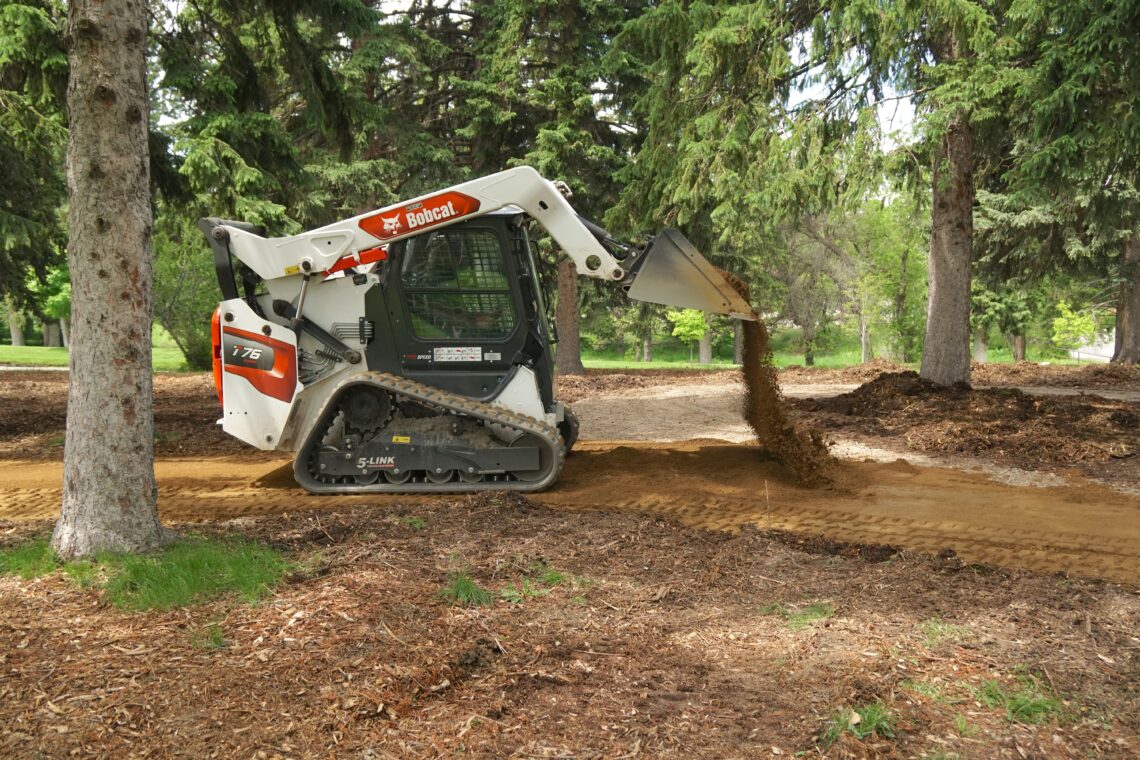 Bobcat skid-steer loader dumping soil in a wooded park, dirt pouring from bucket onto the ground amidst trees and mulch-covered soil.