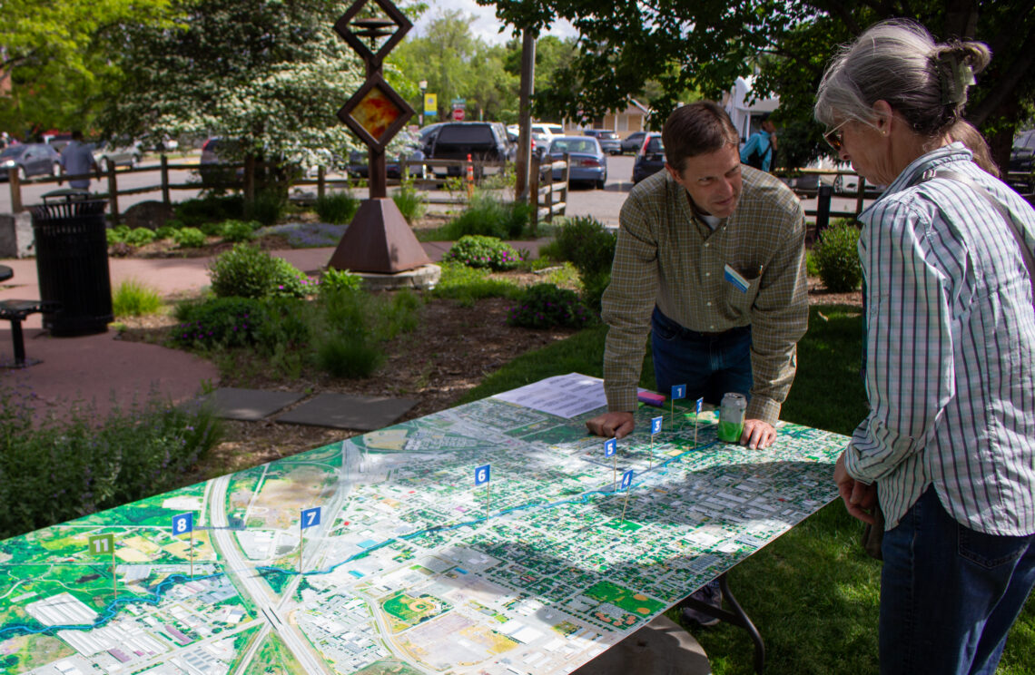 Two adults study a large outdoor map spread on a table, leaning in and pointing at numbered markers.