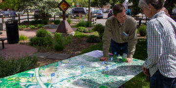 Two adults study a large outdoor map spread on a table, leaning in and pointing at numbered markers.