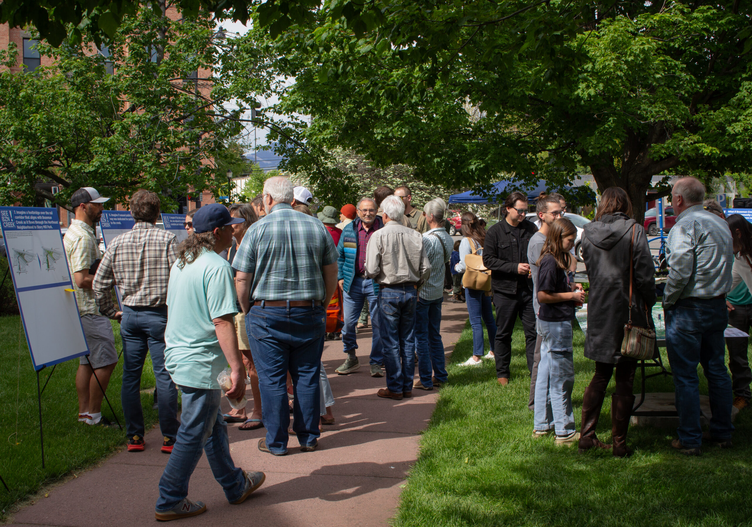 Group of people gathered at an outdoor event along a path, reading informational posters under trees.