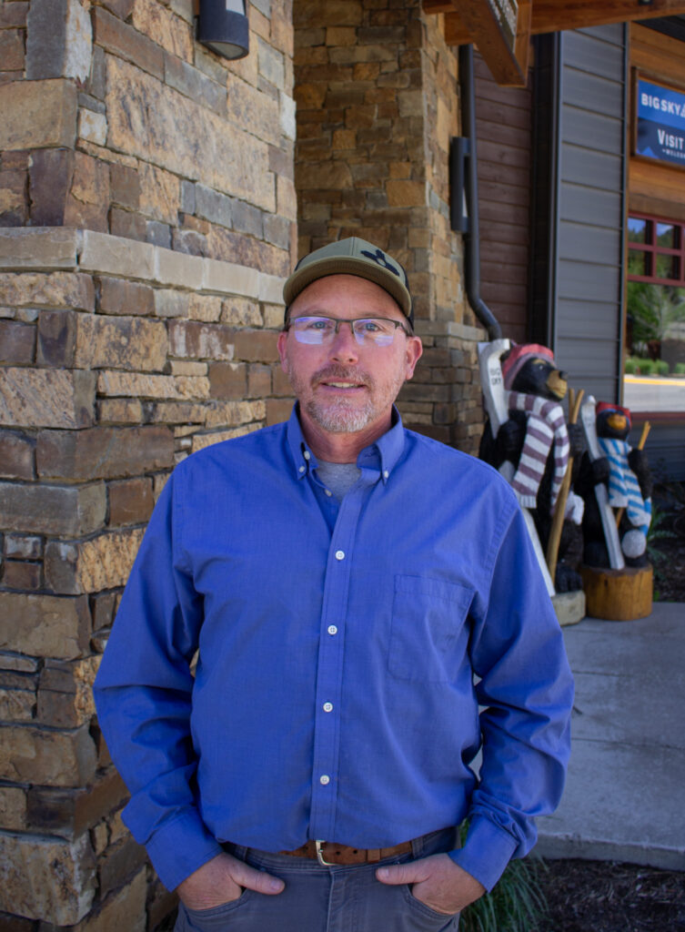 Man in blue button-down shirt and cap stands in front of a stone wall outside a wooden building, hands in pockets.