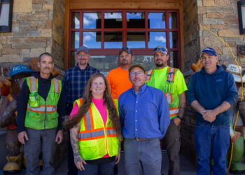 Group of seven construction workers in safety vests posing in front of a stone building entrance.