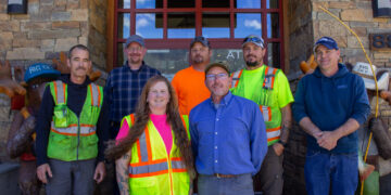 Group of seven construction workers in safety vests posing in front of a stone building entrance.