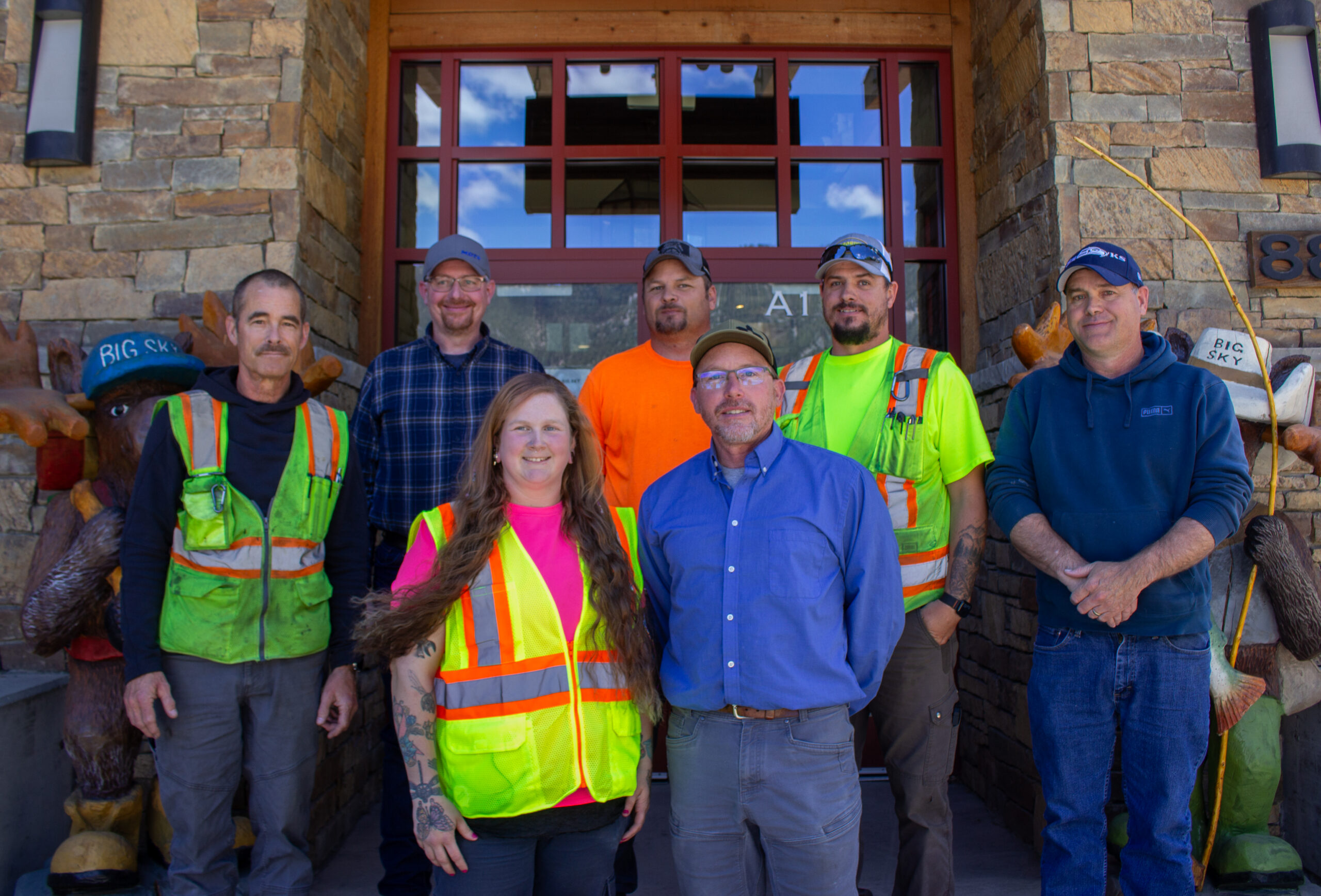 Group of seven construction workers in safety vests posing in front of a stone building entrance.
