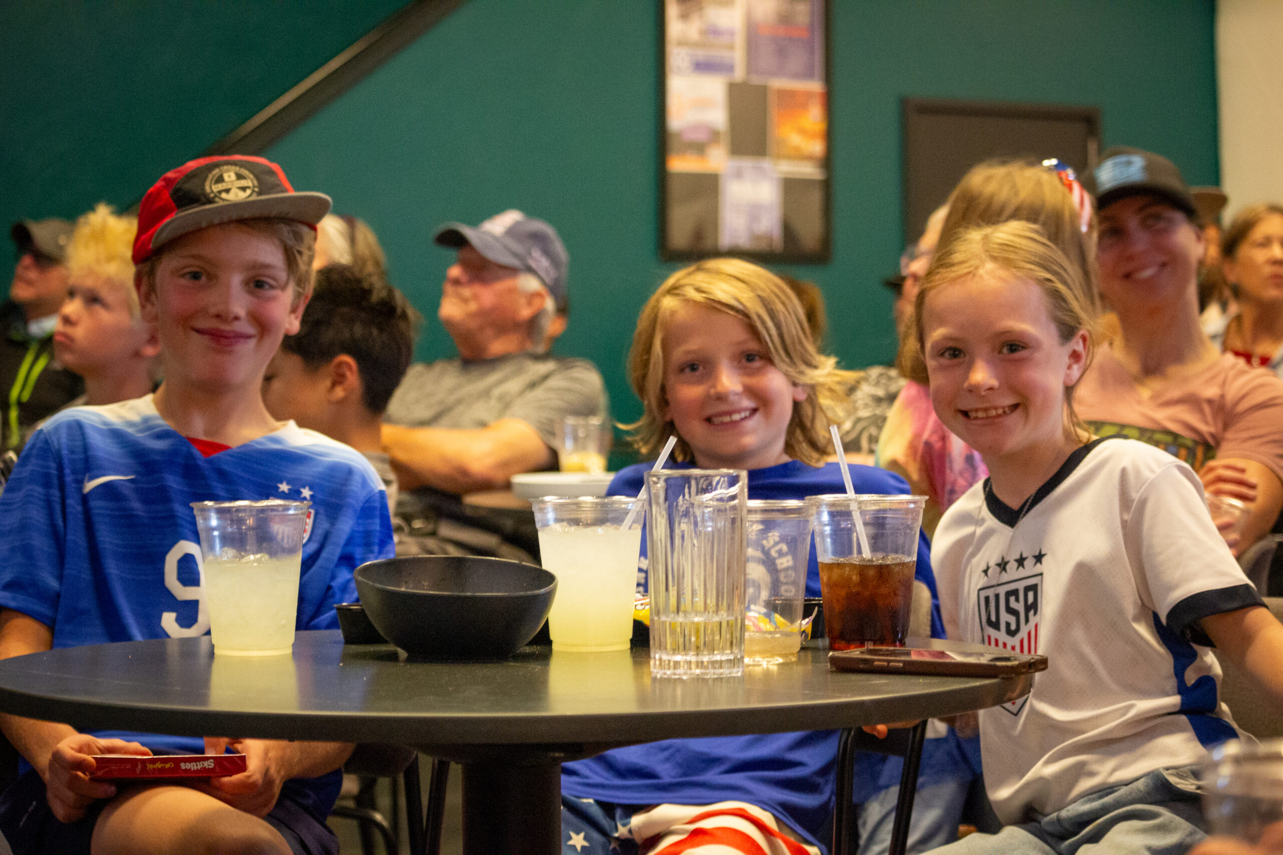 Three smiling kids sit at a round table with drinks and snacks indoors, facing the camera of a casual gathering.