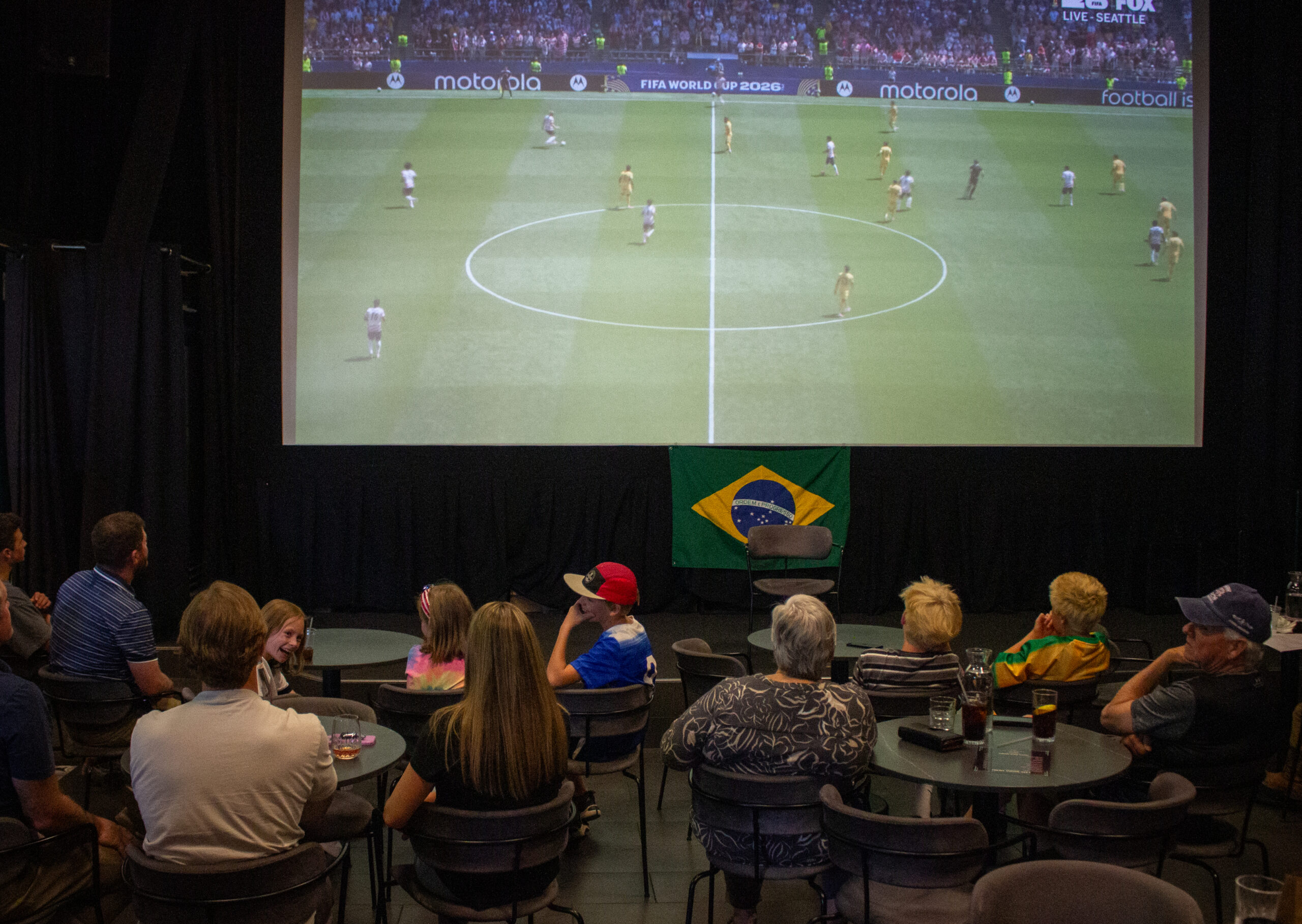 Audience seated at round tables watching a large-screen projection of a soccer match, with a Brazil flag banner beneath the screen.