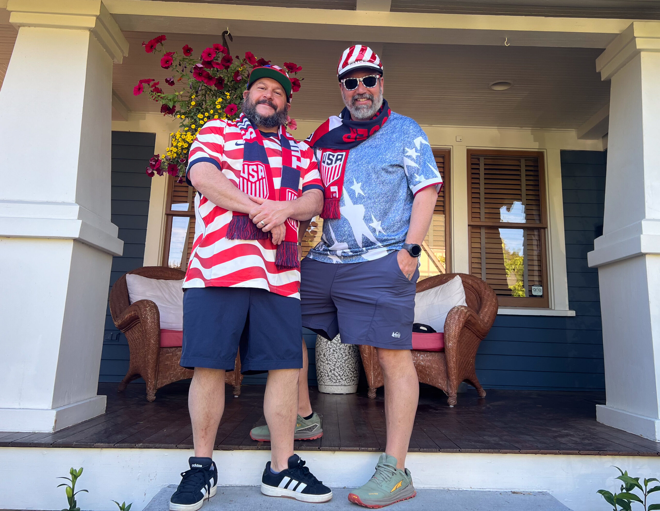 Two men in USA soccer attire pose on a porch, wearing red‑white‑blue jerseys and scarves with a flower-filled backdrop of hanging baskets behind them.