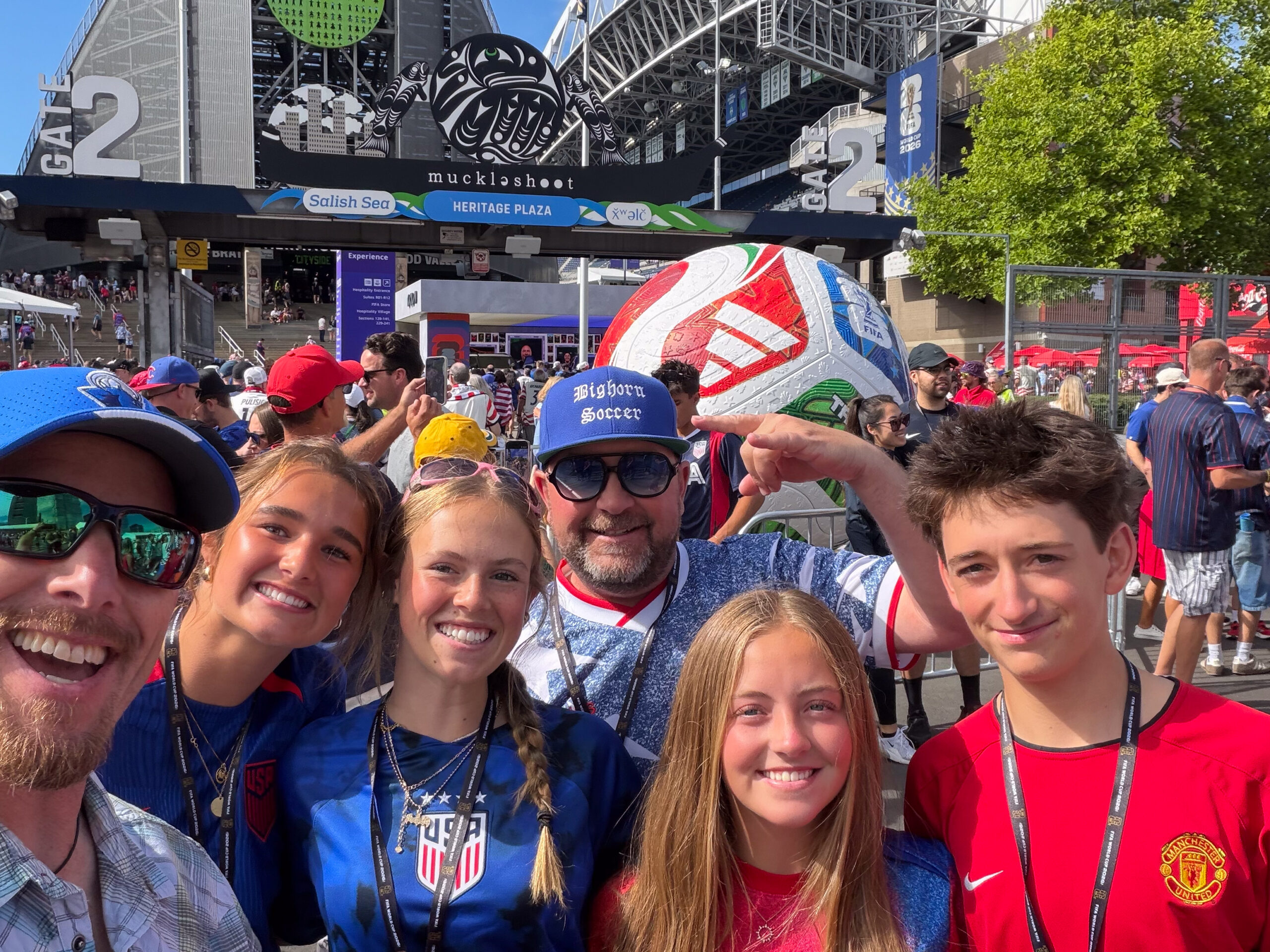 Group of fans taking a selfie at a stadium entrance with a large FIFA World Cup ball sculpture and signage in Heritage Plaza