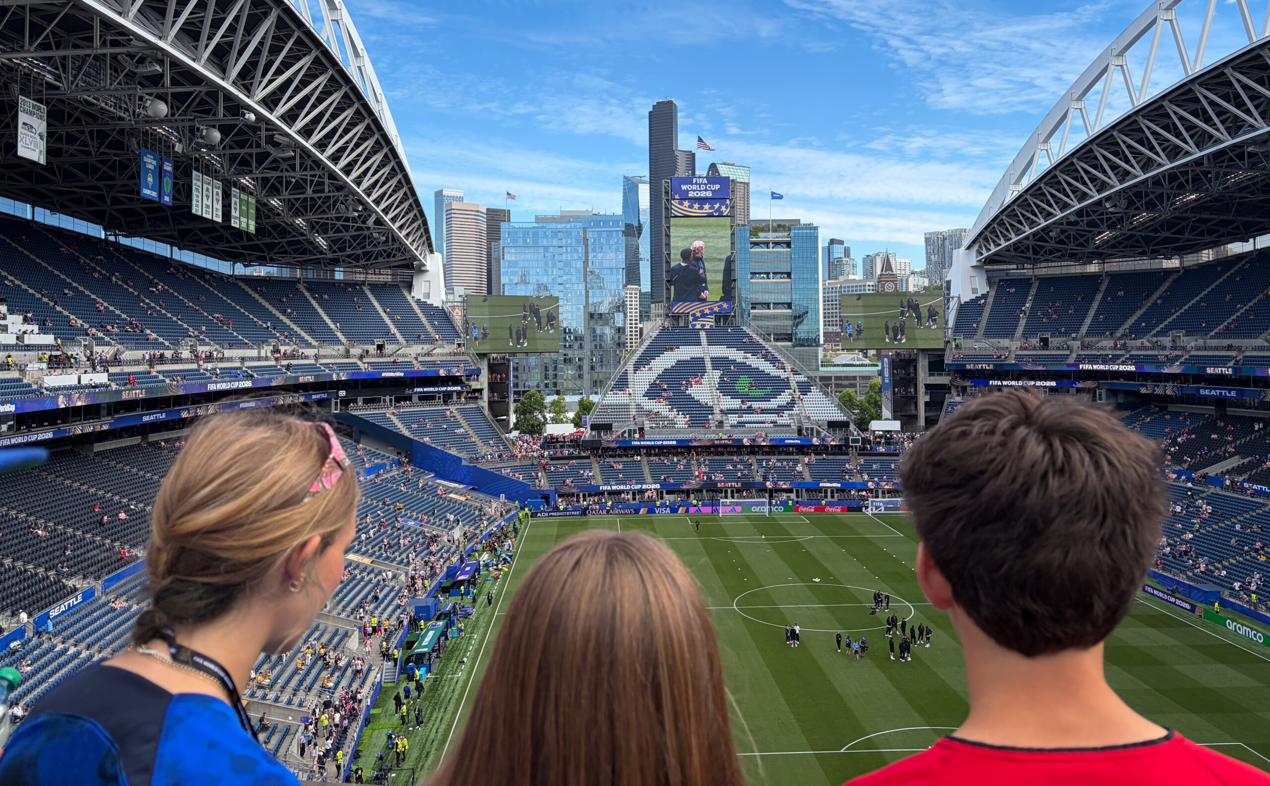 View from the stands inside a large soccer stadium with a city skyline in the distance, huge video screens, and FIFA World Cup 2026 banners visible.