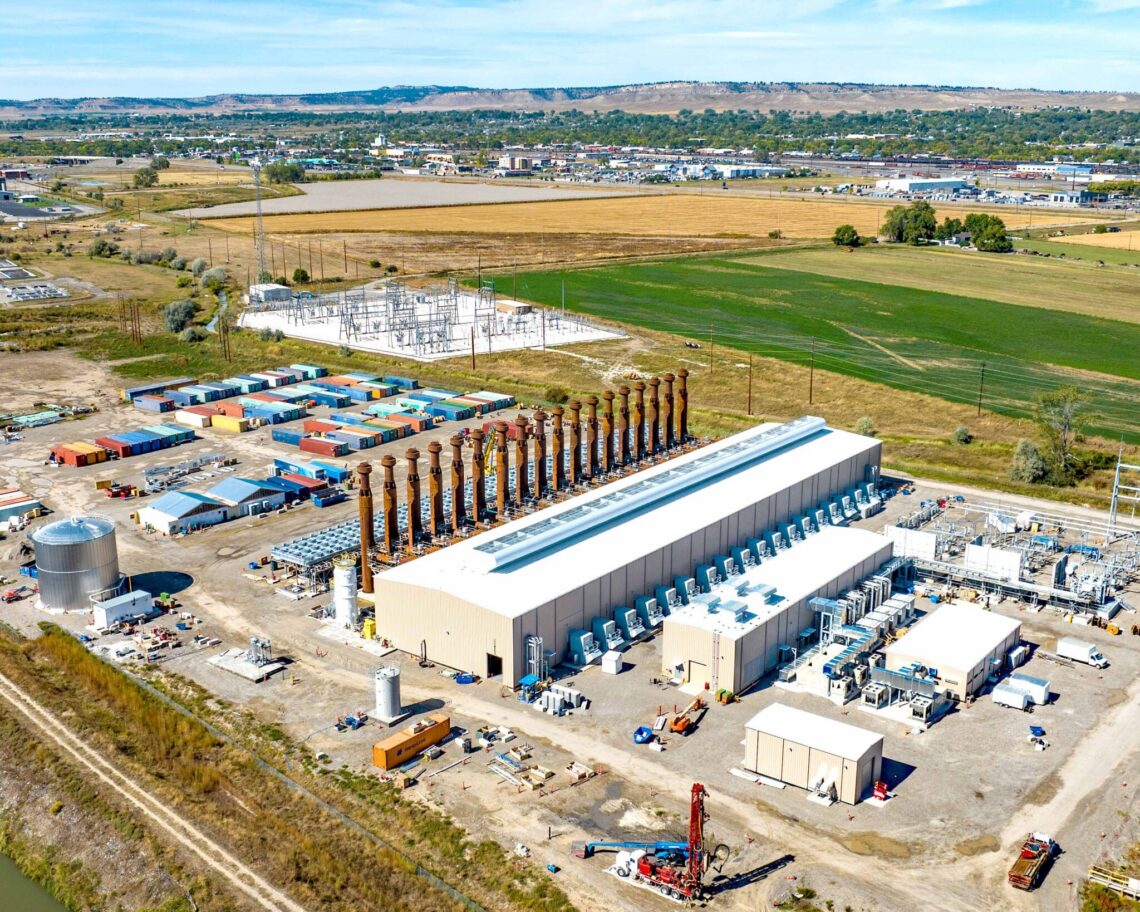 Aerial view of an industrial complex with transformers, equipment yards, and storage containers near open fields and distant town hillsides.
